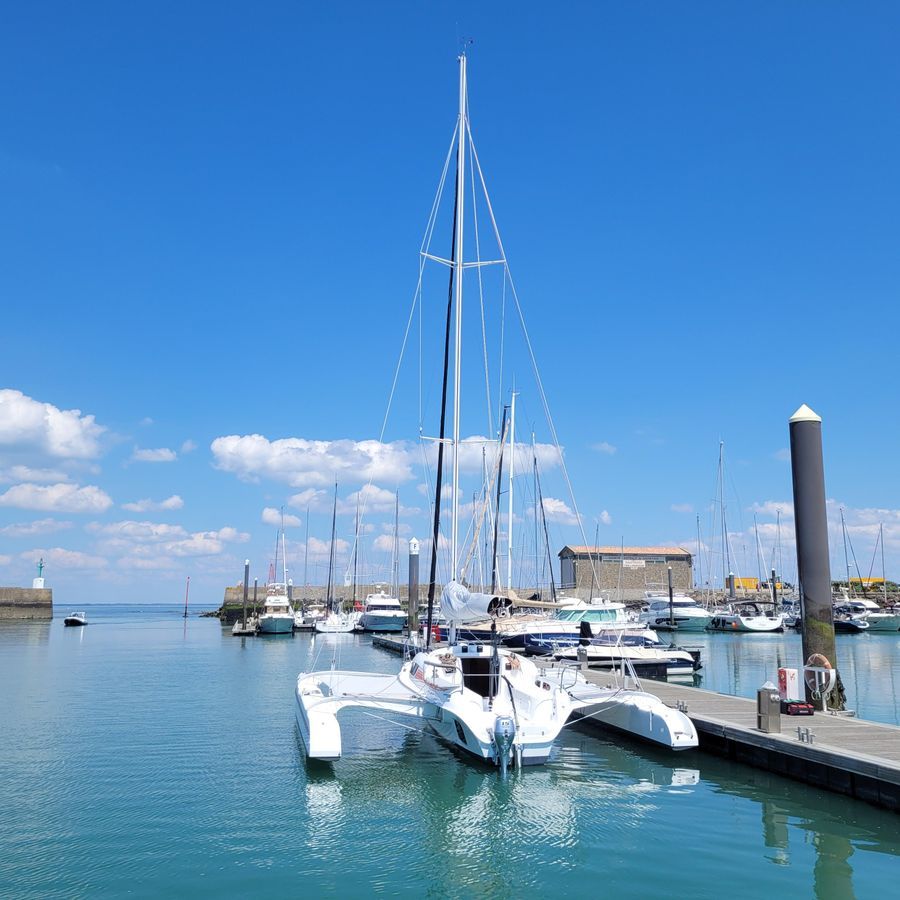 Un trimaran est amarré dans une marina par une journée claire et ensoleillée. D'autres bateaux sont amarrés en arrière-plan avec un ciel bleu et des nuages duveteux en toile de fond.