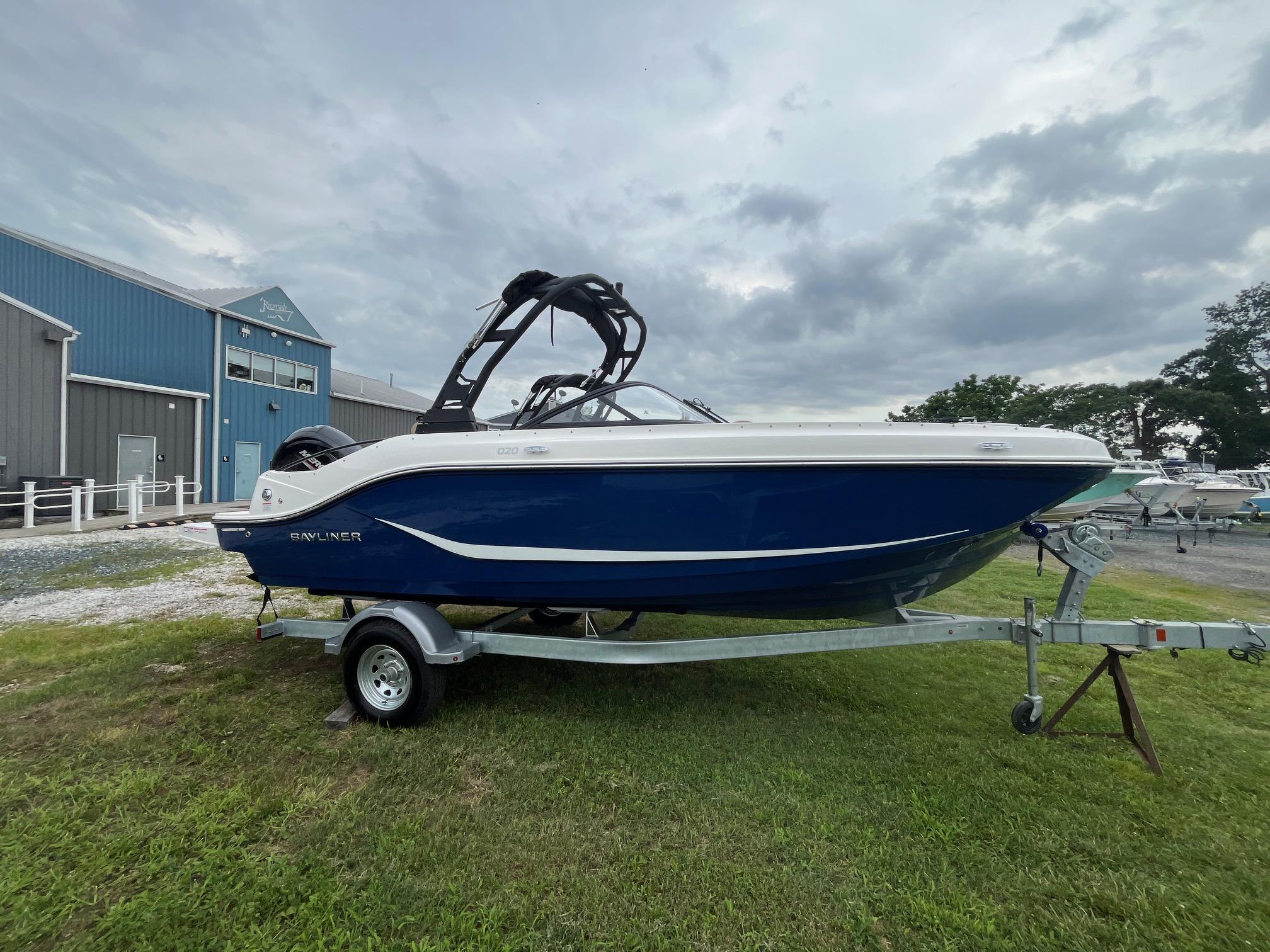 Bayliner D20 2025 Bayliner D20 boat on trailer, parked on grass near blue building under cloudy sky.