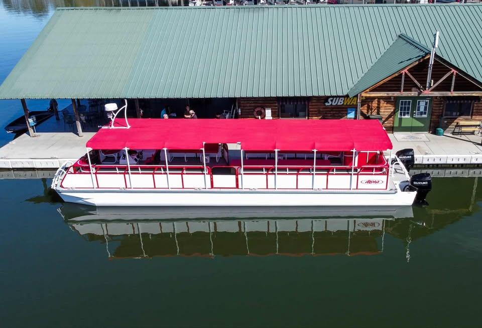 Aloha 50' Water Taxi 2024 Aloha Pontoon 50' Water Taxi with red canopy docked by a lakeside building.