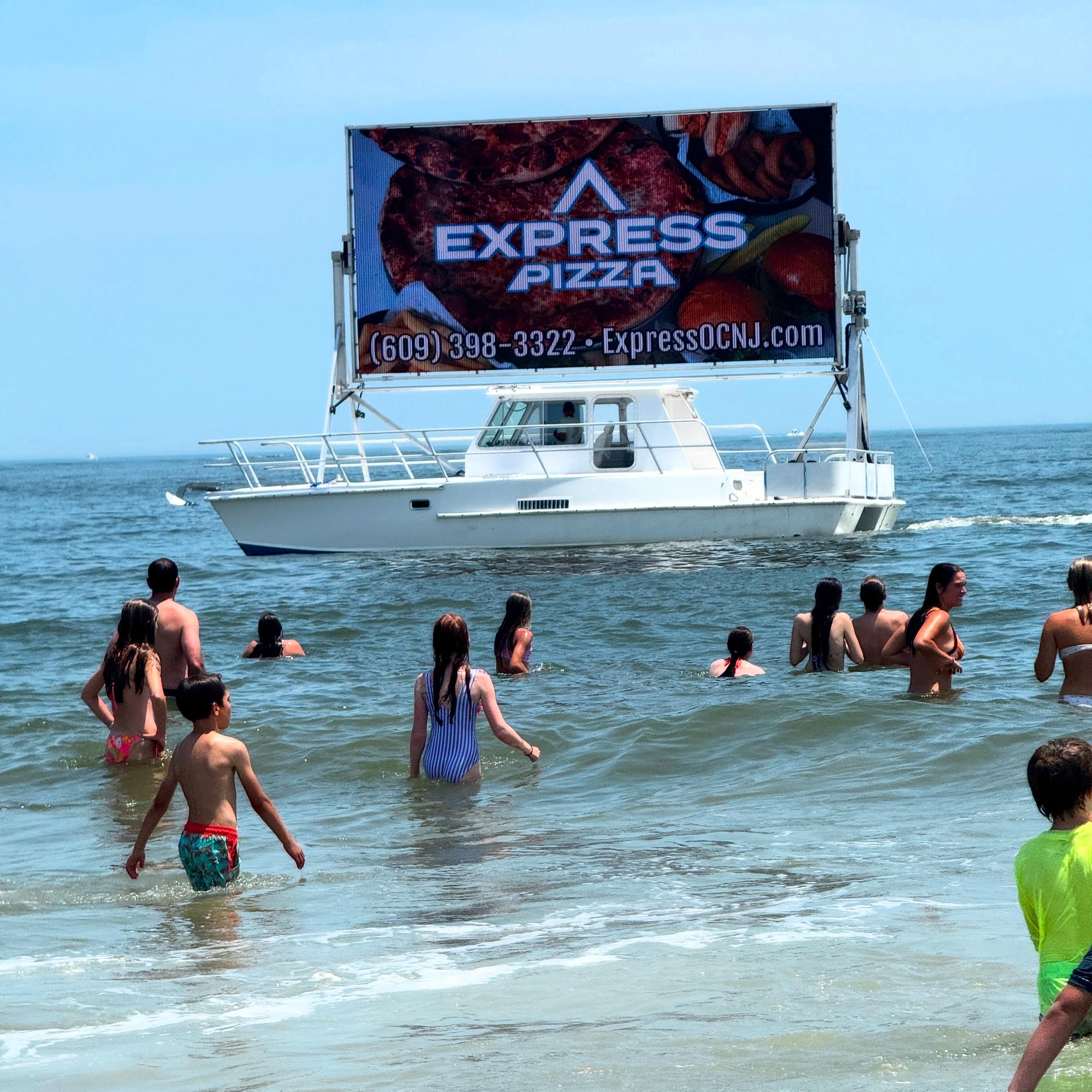 Searunner Custom Cabin Boat with pizza advertisement sails near beachgoers in the ocean.