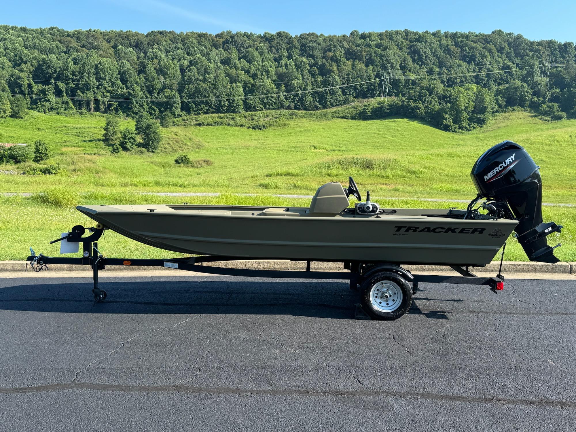 Tracker Grizzly 1648 SC 2025 Tracker Grizzly 1648 SC boat on trailer, parked on asphalt with green hills in background.