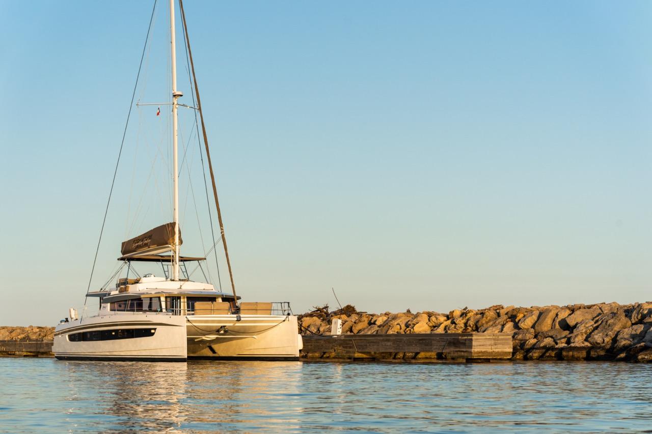 Bali 5.4. Luxurious 2022 Bali 5.4 catamaran docked by rocky shoreline under clear blue sky.