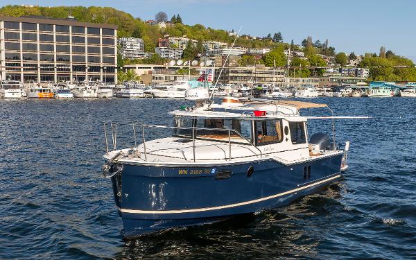 Ranger Tugs R-27 2020 Ranger Tugs R-27 boat on a scenic waterfront with buildings and greenery.