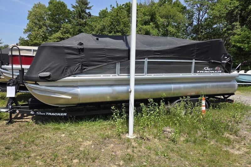 Sun Tracker PB 18 DLX 2025 Sun Tracker PB 22 RF pontoon boat covered, parked on trailer, surrounded by greenery.