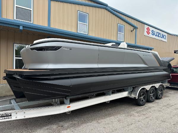 Manitou Explore Navigator Dual Engine Rotax 2024 Manitou Explore Navigator Dual Engine Rotax pontoon boat on a trailer outside a Suzuki dealership.