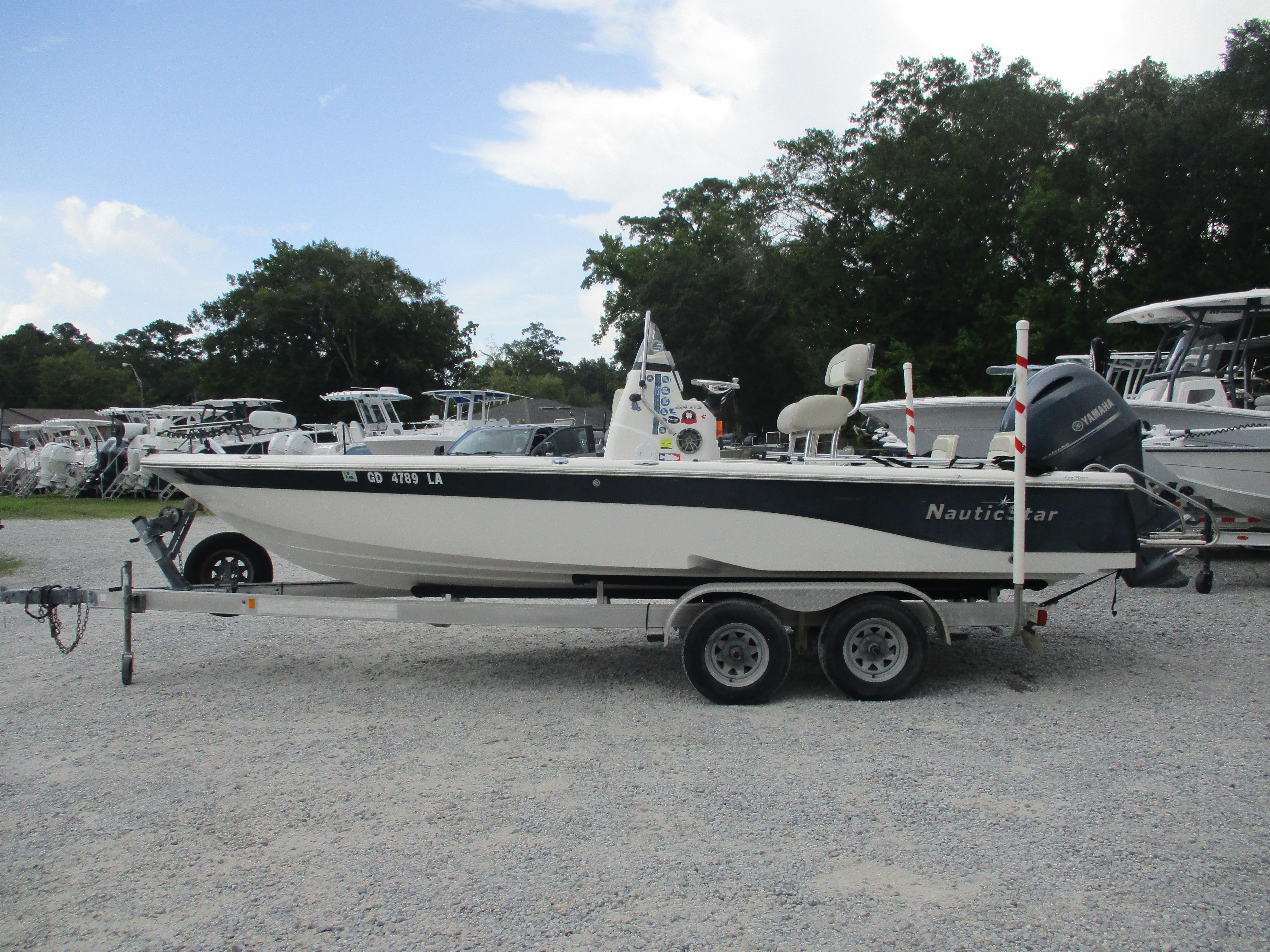 NauticStar 224 2015 NauticStar 224 boat on trailer, parked on gravel, surrounded by trees and other boats.
