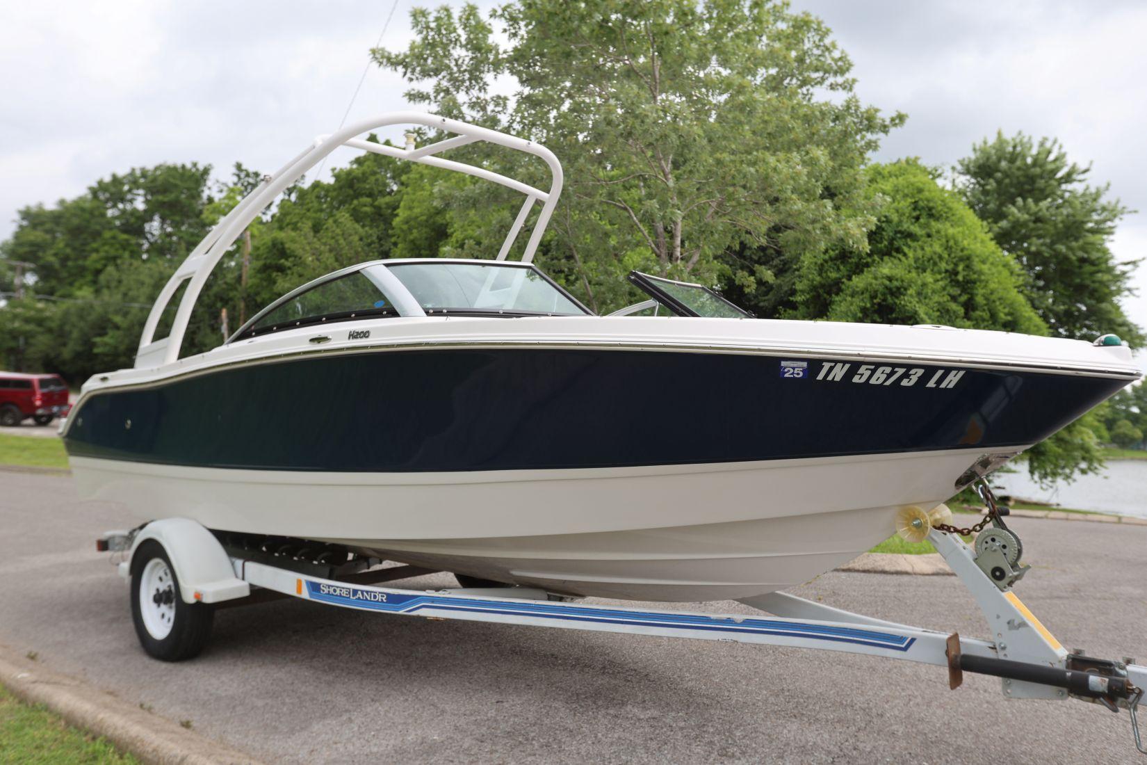 Four Winns H200 2015 Four Winns H200 boat on trailer, parked near a lake with lush greenery.