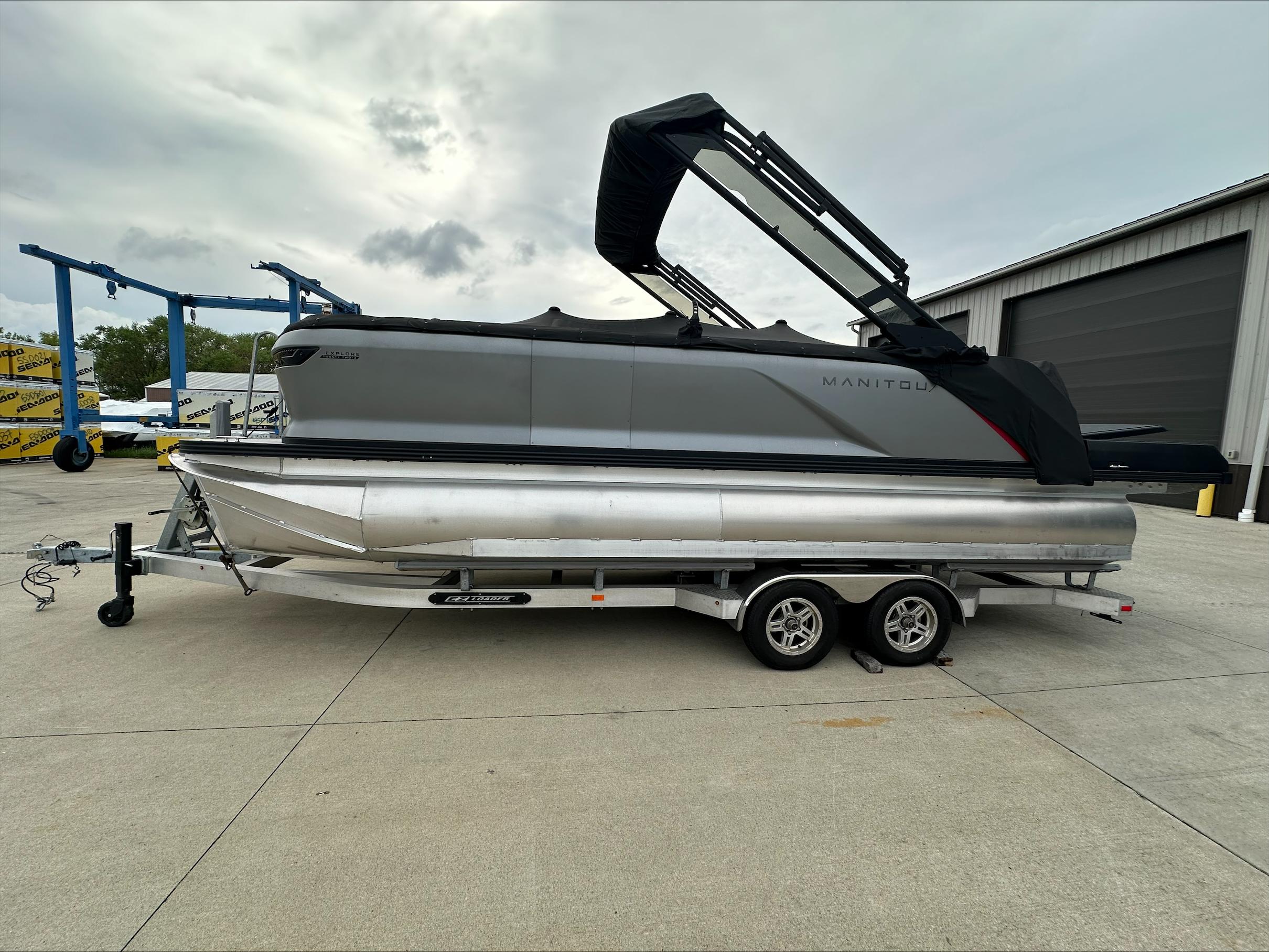 Manitou Explore 2023 Manitou Explore pontoon boat on trailer, parked outdoors near a warehouse.
