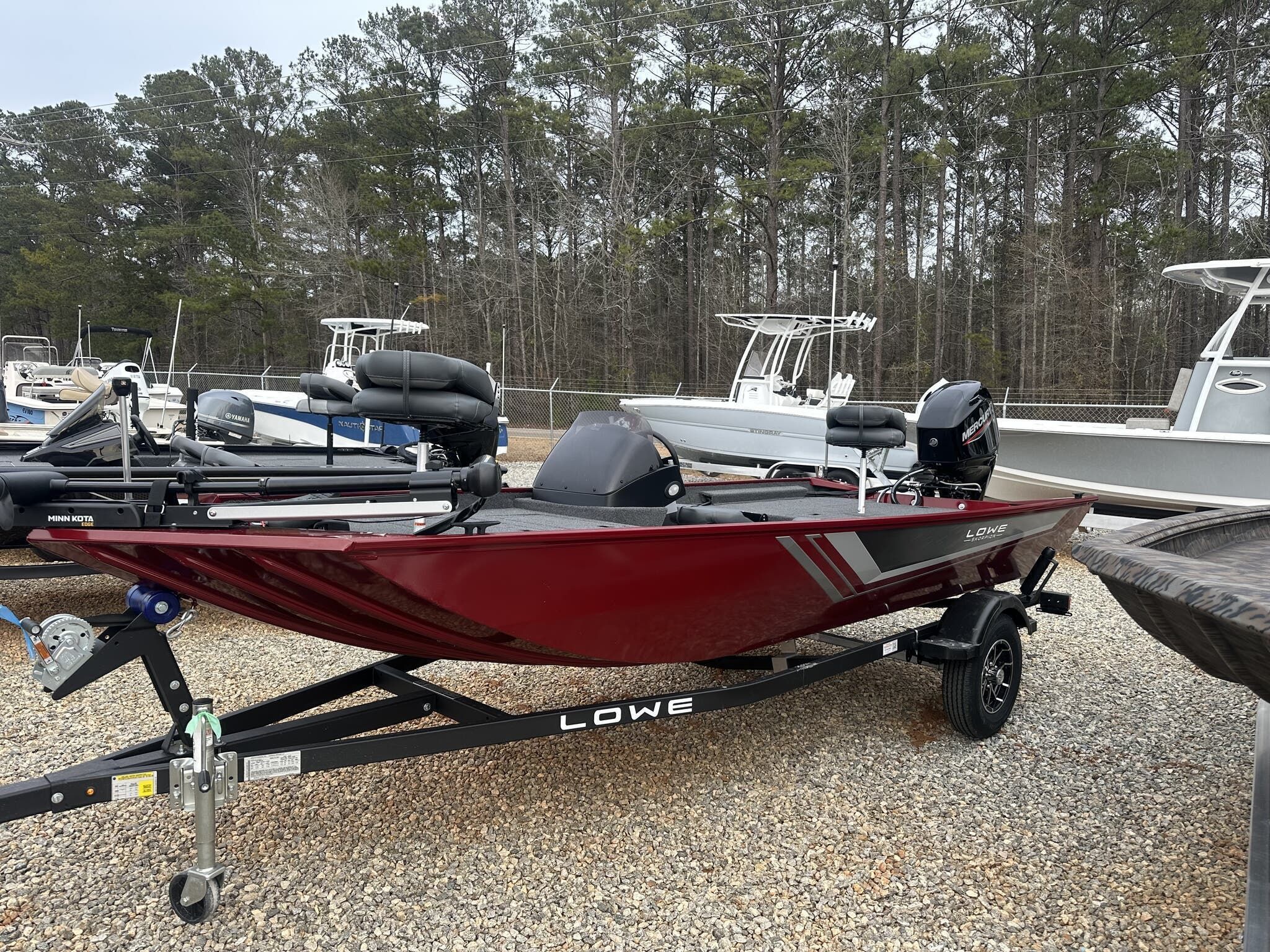 Lowe Skorpion 2025 Lowe Skorpion boat in red, parked on a gravel lot with trees in the background.