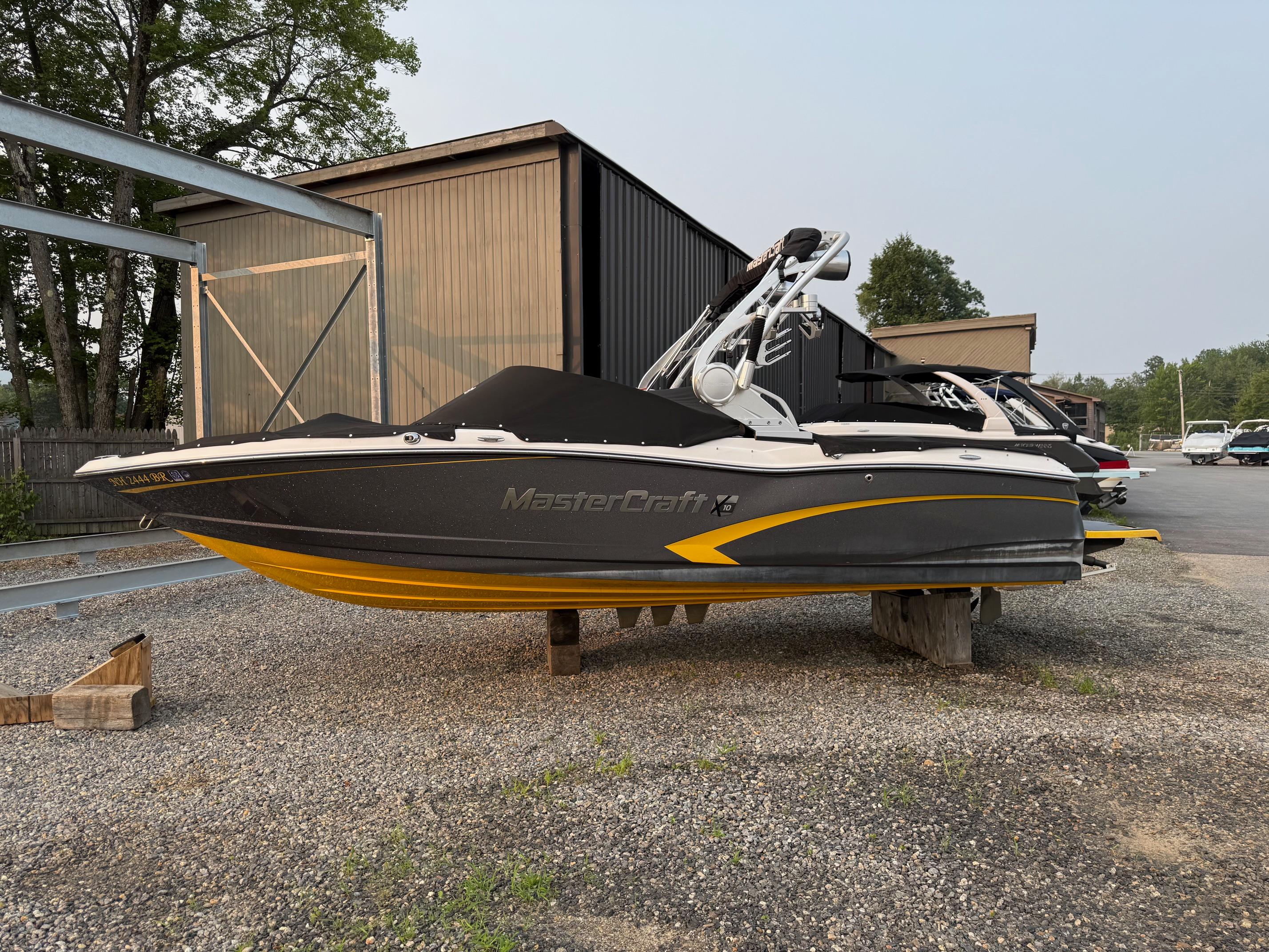 MasterCraft X10 2015 MasterCraft X10 boat with black cover, parked on gravel near storage buildings.