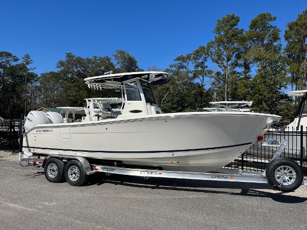 Cobia 265 Center Console 2025 Cobia 265 Center Console boat on trailer, parked outdoors with trees in background.
