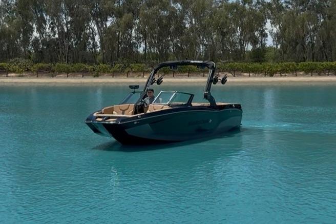 MasterCraft XT22 2025 MasterCraft XT22 boat cruising on clear blue water near a tree-lined shore.