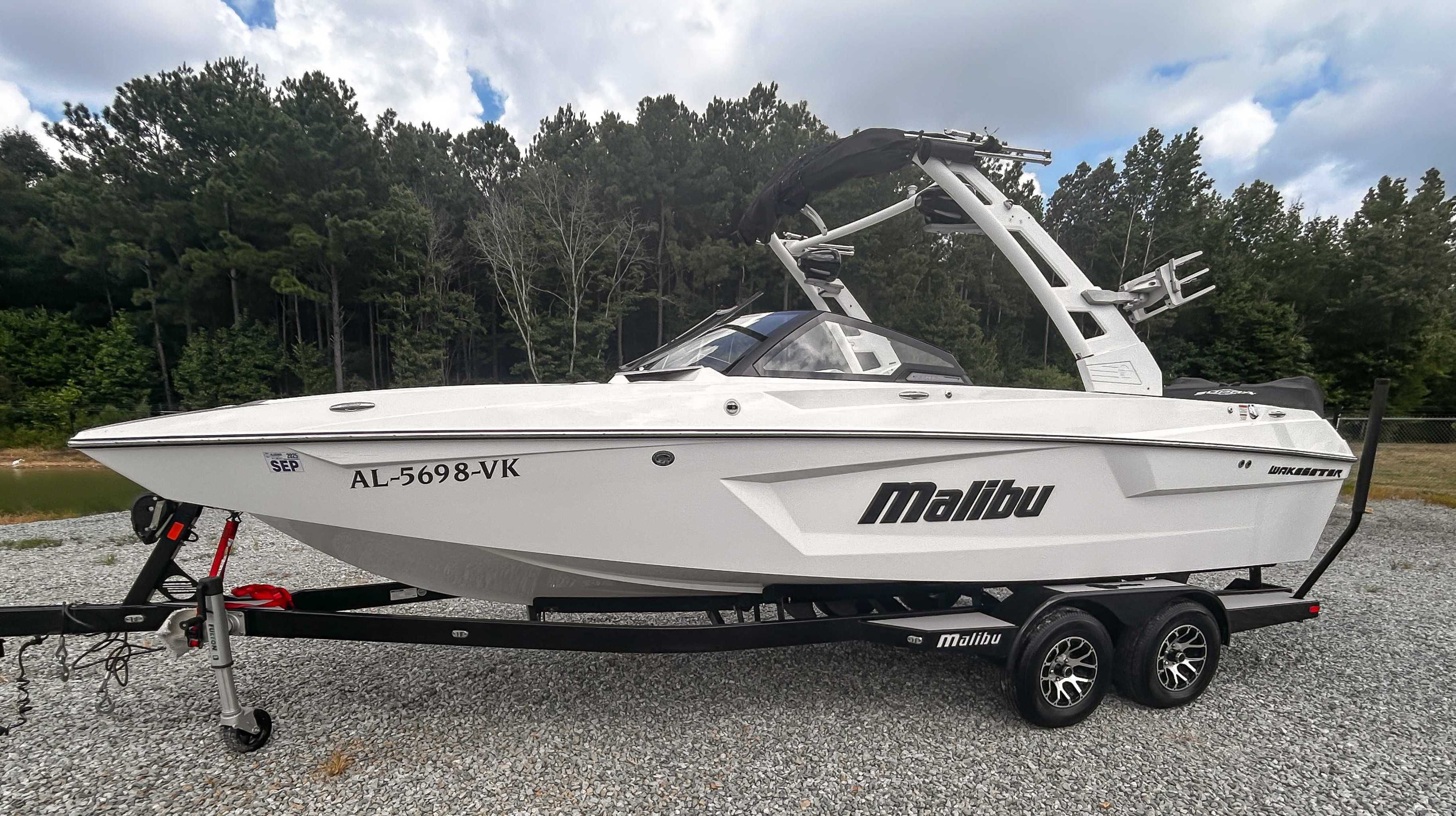 Malibu 23 MXZ 2020 Malibu 23 MXZ boat on trailer, parked on gravel, surrounded by trees.