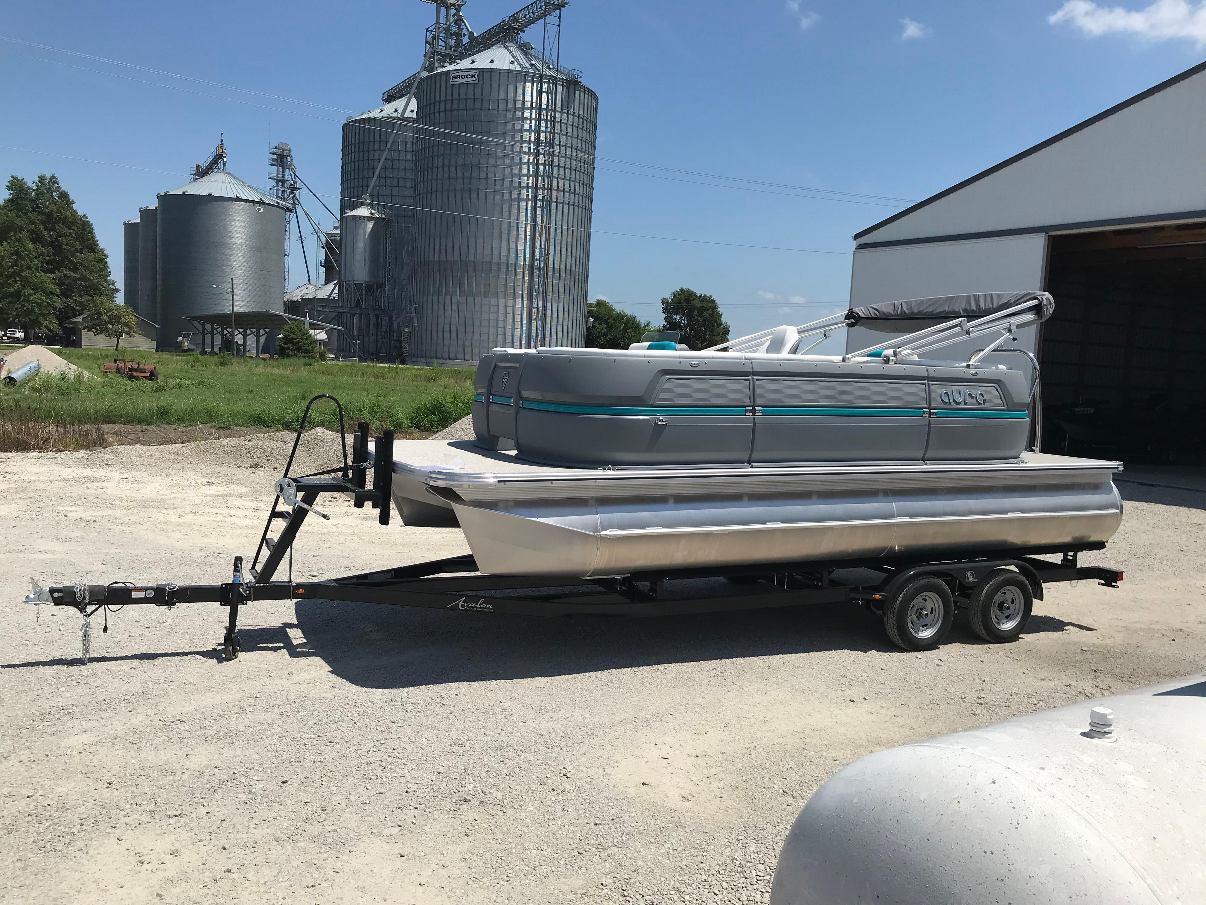 Avalon AURA 2025 Avalon AURA pontoon boat on trailer, parked near industrial silos and a barn.