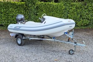 AB Inflatables Rider 2024 AB Inflatables Rider boat on trailer, parked on gravel with greenery background.