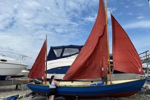 Drascombe Longboat 1970 Drascombe Longboat with red sails on a trailer, parked beside other boats.
