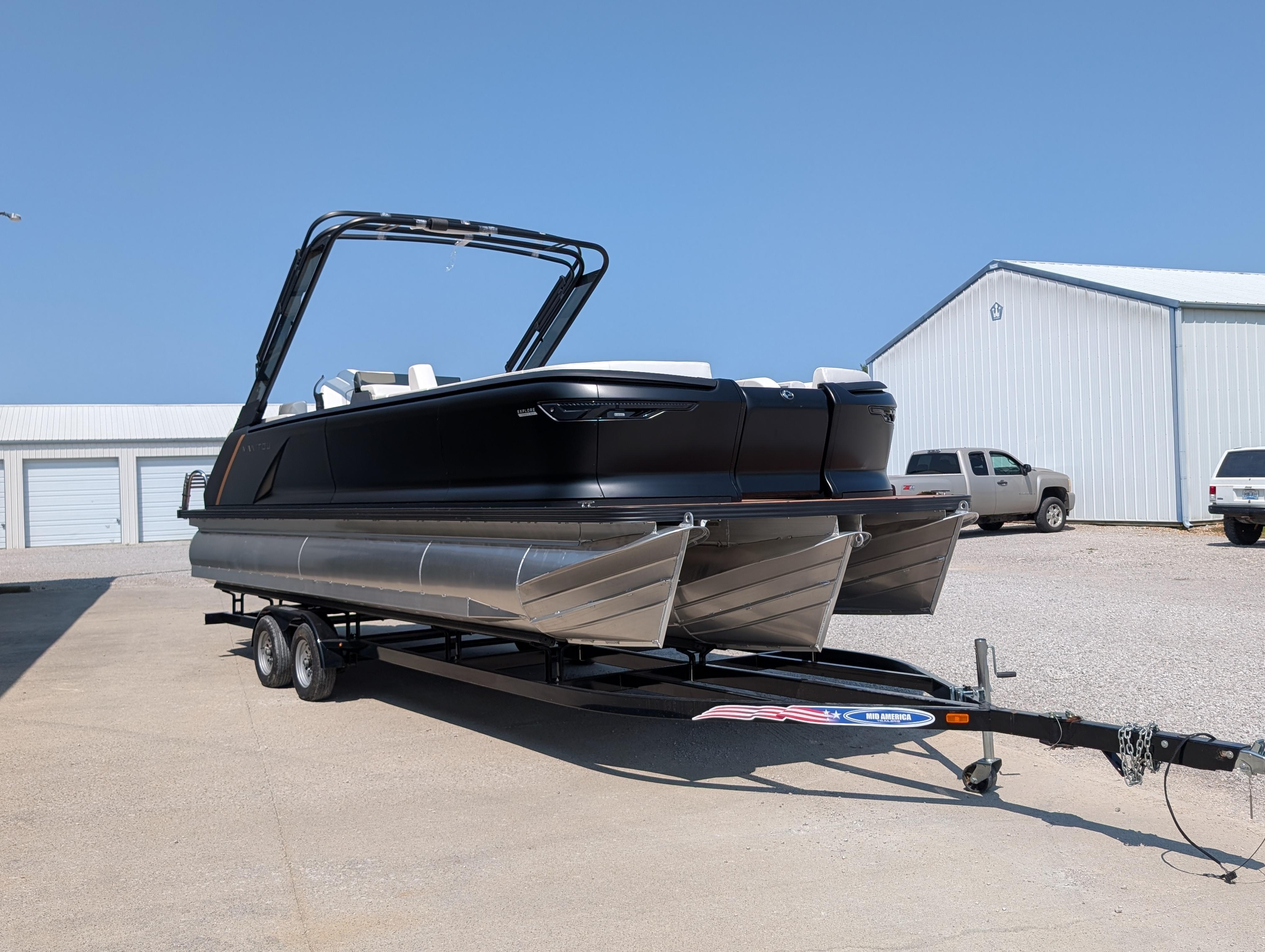 Manitou Explore 2025 Manitou Explore pontoon boat on trailer, parked near storage buildings under clear blue sky.