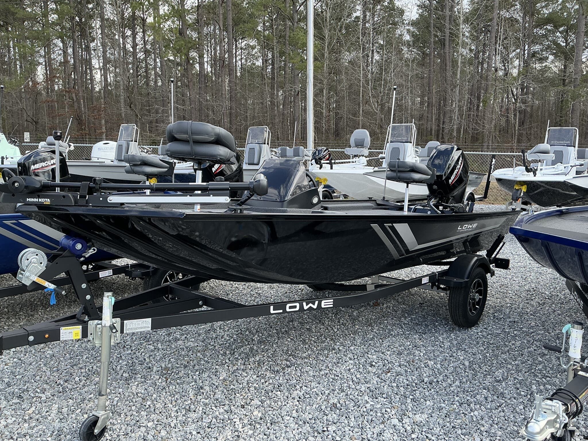 Lowe Skorpion 2025 Lowe Skorpion boat on trailer, parked on gravel, surrounded by trees and other boats.