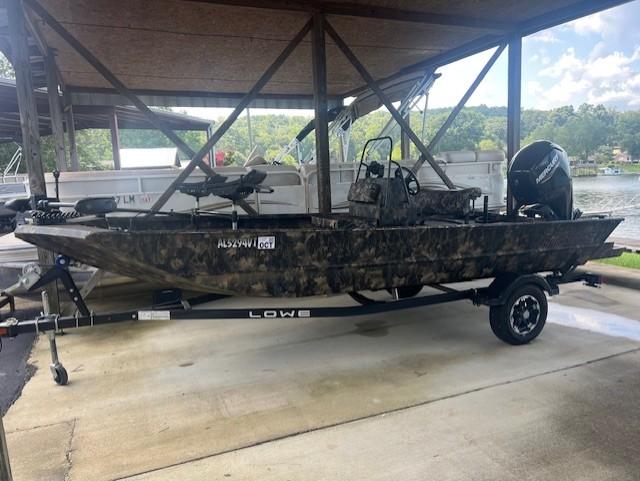 Lowe Roughneck 1860 CC 2023 Lowe Roughneck 1860 CC boat on trailer under shelter, camo design, parked near lake.