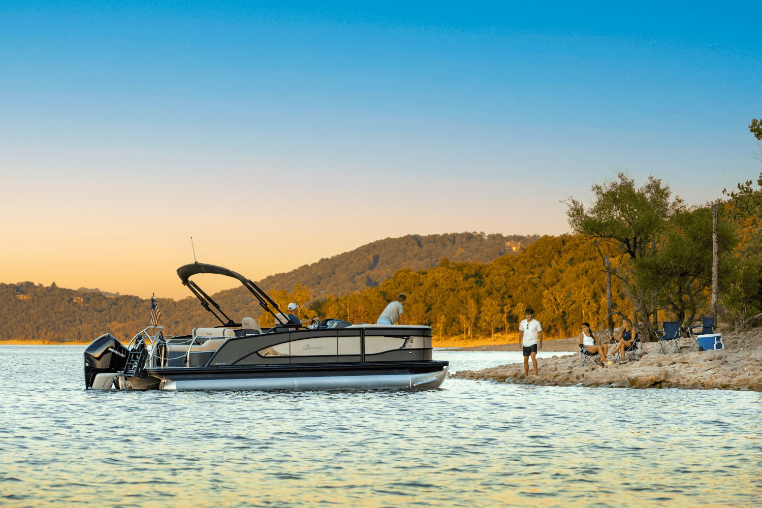 Barletta Lusso 23 UC Barletta Lusso 23 UC 2026 pontoon boat near scenic lakeside with people relaxing.