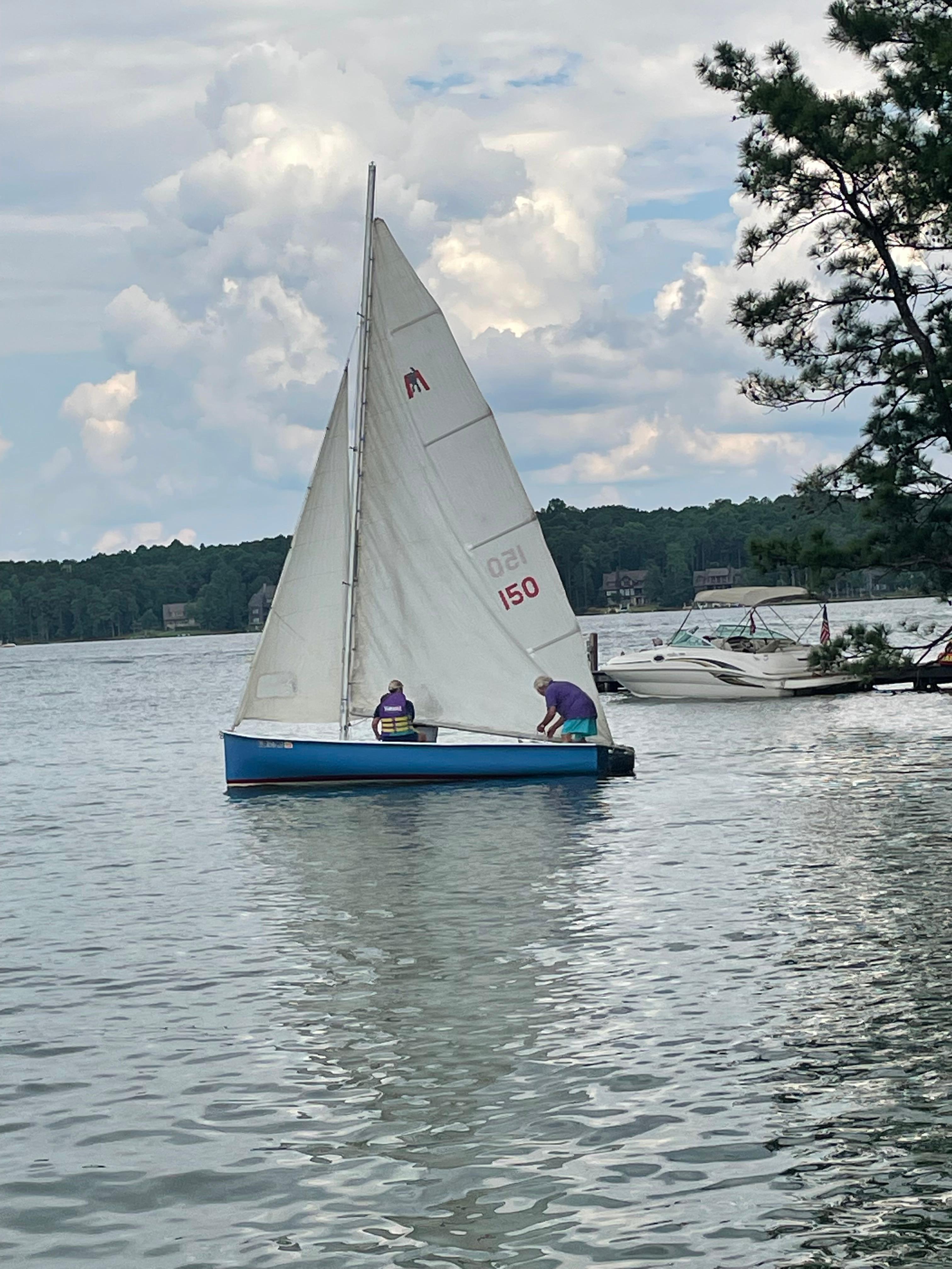 Sailboat MobJack Sailboat MobJack 1965 on a calm lake with cloudy sky and nearby trees.