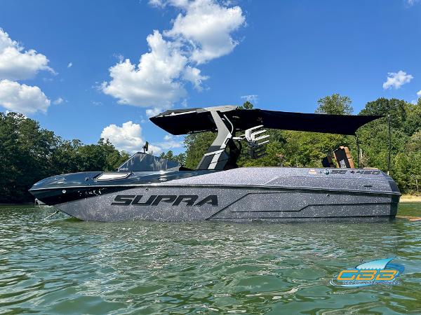 Supra SE 2024 Supra SE boat on a lake, under a clear blue sky with trees in the background.