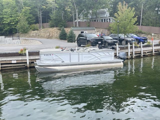 Sylvan 8522 LZ 2022 Sylvan 8522 LZ pontoon boat docked by a lakeside with parked cars nearby.